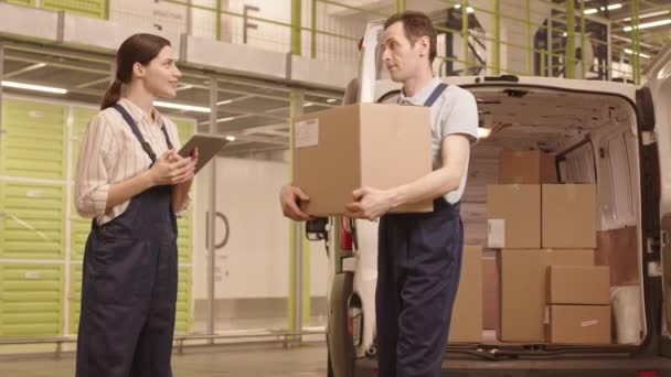 Средний белый мужчина разгружает картонную коробку из машины доставки, показывая на ней ярлык женщине-менеджеру в комбинезоне, стоящей на складе и использующей планшетный компьютер, они разговаривают - Кадры, видео