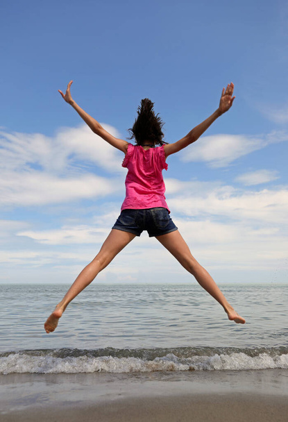 młoda dziewczyna skacze bardzo wysoko z otwartymi ramionami i nogami nad morzem w lecie - Zdjęcie, obraz