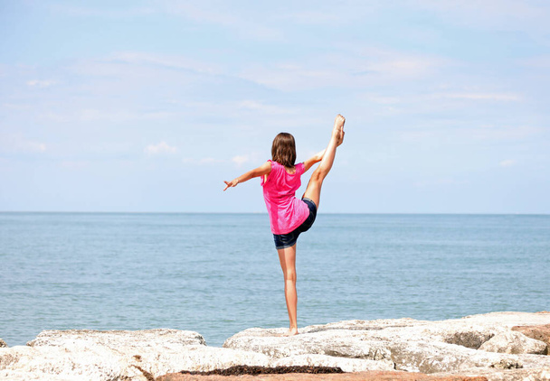 kaukaska dziewczyna wykonywania gimnastyki rytmicznej ćwiczenia z nogą w górę i stóp nad głową nad morzem w lecie - Zdjęcie, obraz