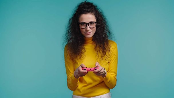 KYIV, UKRAINE - OCTOBER 1, 2021: brunette young woman in glasses holding joystick and playing isolated on blue - Fotoğraf, Görsel
