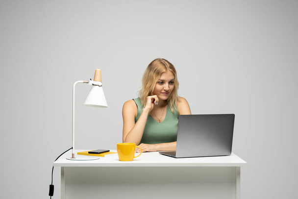 Smiling business woman working with a laptop isolated on a grey background. Portrait of a pretty young woman studying while sitting at the table with grey laptop computer, notebook. - Zdjęcie, obraz