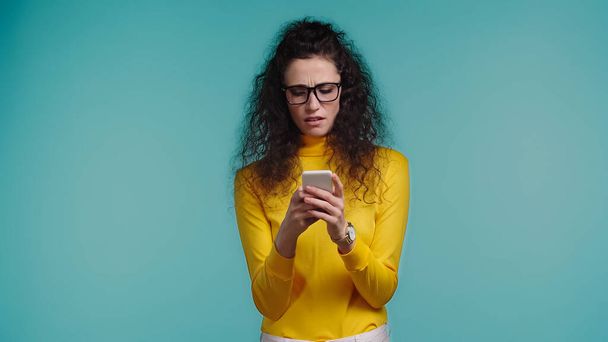 displeased young woman in glasses texting on smartphone isolated on blue  - Foto, imagen