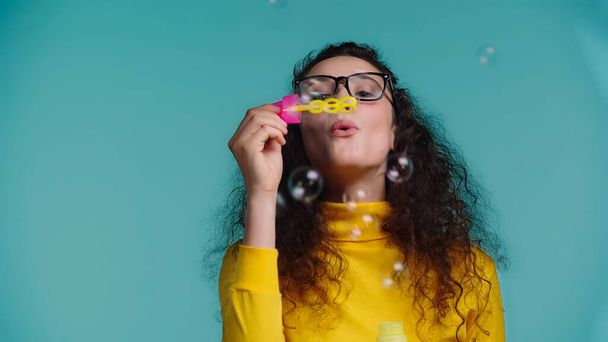 young woman in glasses blowing soap bubbles isolated on blue - Foto, Bild