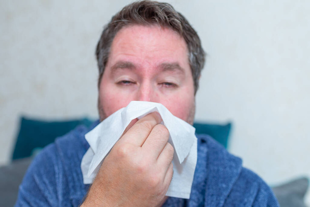 healthcare, people and medicine concept - ill man blowing nose at home. sick male at home have health problems, get flu or fever symptoms, tired guy suffer from sickness sneezing holding tissue. - Foto, imagen