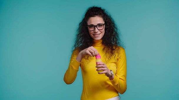 happy young woman in glasses holding bottle with soap bubbles isolated on blue - Фото, изображение