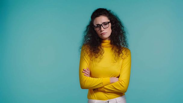 displeased young woman in glasses and turtleneck standing with crossed arms isolated on blue - Fotoğraf, Görsel