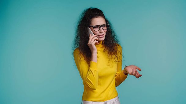 displeased young woman in glasses talking on smartphone isolated on blue  - Fotografie, Obrázek