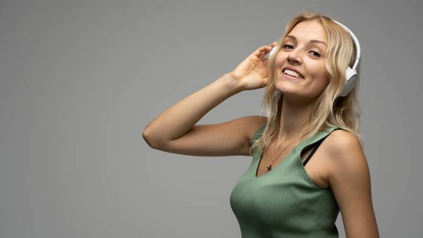Beautiful attractive young blond woman wearing green t-shirt and glasses in white headphones listening music and smiling on grey background in studio. Relaxing and enjoying. Lifestyle. - Zdjęcie, obraz