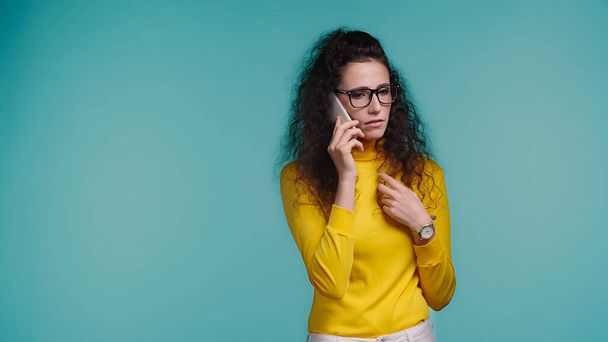curly young woman in glasses talking on smartphone isolated on blue  - Fotó, kép