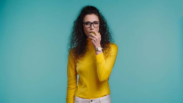 young curly woman eating apple isolated on blue   - Foto, imagen