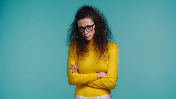 offended young woman in glasses and turtleneck standing with crossed arms isolated on blue  - Fotografie, Obrázek