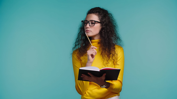 thoughtful young woman in glasses and turtleneck holding notebook and pen isolated on blue  - Foto, imagen