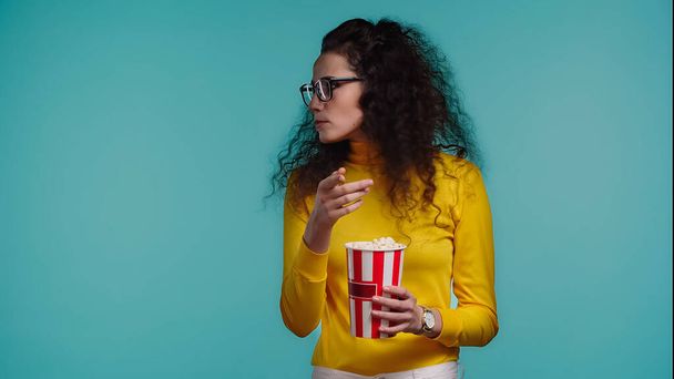 curly young woman holding popcorn bucket isolated on turquoise  - Foto, afbeelding