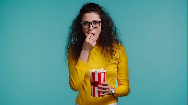 curly young woman holding bucket while eating popcorn isolated on blue - Fotoğraf, Görsel