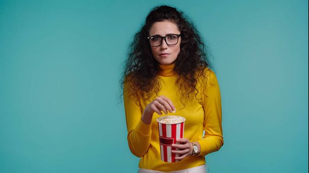 lockige junge Frau schaut Film und hält Popcorn-Eimer isoliert auf blauem Grund   - Foto, Bild