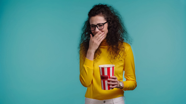 счастливая молодая женщина держит ведро попкорна и смотрит комедийный фильм изолированы на синий   - Фото, изображение