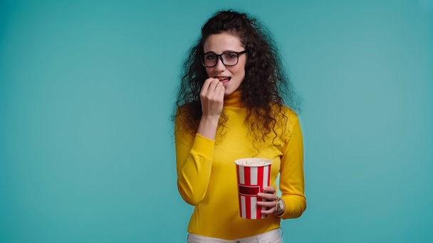 kudrnatá mladá žena jíst popcorn při sledování filmu izolované na modré   - Fotografie, Obrázek