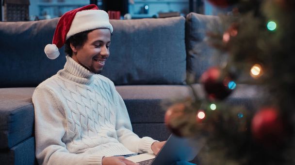 サンタ・ハットの陽気なアフリカ系アメリカ人男性がクリスマスツリーの近くにノートパソコンを使って - 写真・画像