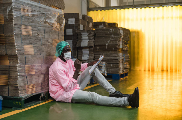 Gerenciador de controle de inventário masculino africano em assento uniforme estéril e ordem de verificação do pacote de caixa de papelão empilhado em armazém na fábrica de processamento de bebidas - Foto, Imagem