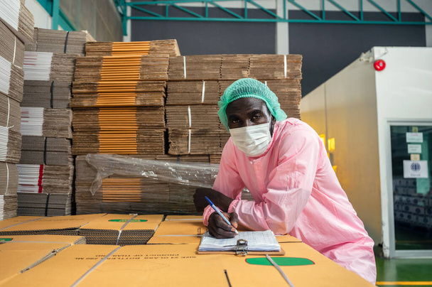 Διευθυντής ελέγχου απογραφής αφρικανού ανθρώπου σε αποστειρωμένο πακέτο ελέγχου ενδυμάτων σε χάρτινη συσκευασία στοιβαγμένη σε αποθήκη σε μονάδα επεξεργασίας ποτών - Φωτογραφία, εικόνα