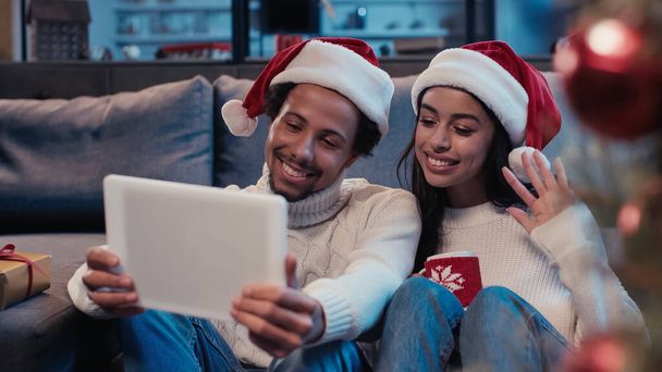 ビデオ通話中に手を振ってデジタルタブレットを見ている女性の近くに幸せなアフリカ系アメリカ人男性 - 写真・画像