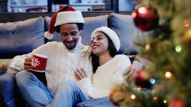 Africano americano hombre sosteniendo taza y sonriendo con novia cerca de árbol de Navidad - Foto, imagen