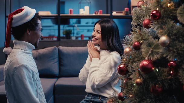 cheerful african american woman smiling while looking at boyfriend in santa hat near christmas tree - 写真・画像