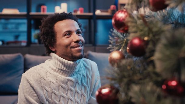 幸せなアフリカ系アメリカ人男性がクリスマスツリーを見ながらセーターを着て - 写真・画像