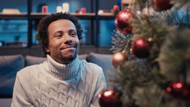 クリスマスツリーを見ながらセーターを着たアフリカ系アメリカ人男性が - 写真・画像