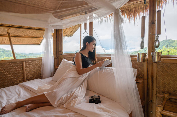 Hermosa joven asiática mujer descansando y leyendo un libro en la cama en choza de paja entre la montaña de vacaciones - Foto, Imagen