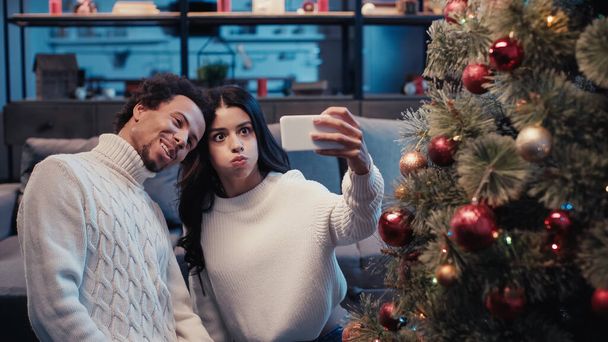 happy african american man taking selfie with woman puffing cheeks near christmas tree - Photo, Image