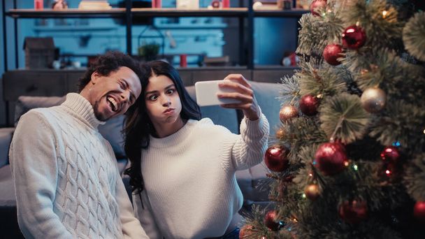 šťastný africký Američan muž přičemž selfie s ženou foukání tváře v blízkosti vánočního stromu - Fotografie, Obrázek