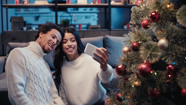 Mutlu Afro-Amerikalı kadın Noel ağacının yanında dilini çıkaran bir adamla selfie çekiyor. - Fotoğraf, Görsel