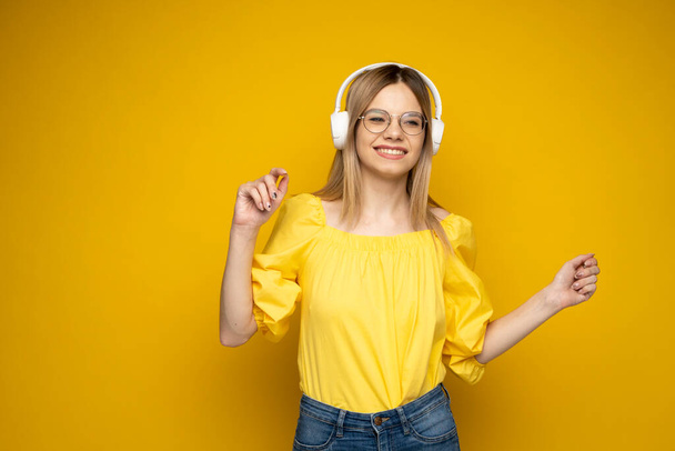 Beautiful attractive young blond woman wearing yellow t-shirt and glasses in white headphones listening music, dancing and laughing on blue background in studio. Relaxing and enjoying. Lifestyle. - Photo, image