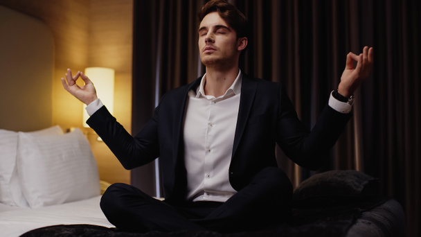young businessman with closed eyes meditating in lotus pose on bed in hotel room - Фото, изображение