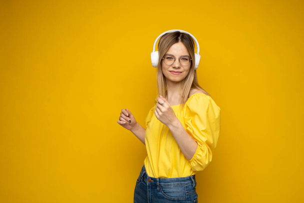 Beautiful attractive young blond woman wearing yellow t-shirt and glasses in white headphones listening music, dancing and laughing on blue background in studio. Relaxing and enjoying. Lifestyle. - Foto, Imagem