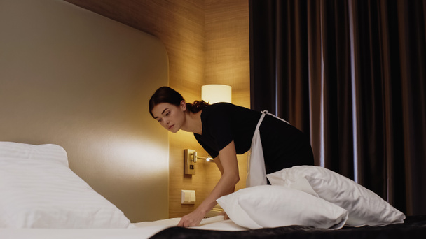 jeune femme de chambre en uniforme et tablier faisant lit dans la chambre d'hôtel - Photo, image