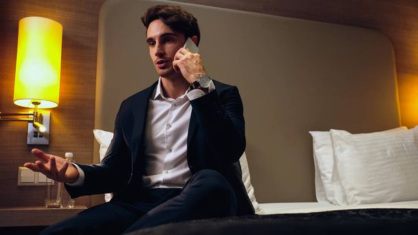 businessman in suit sitting on bed and gesturing while talking on smartphone in hotel room - Foto, afbeelding