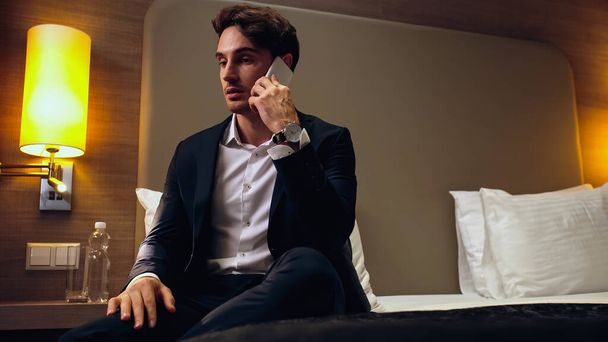 biznesmen siedzi na łóżku i rozmawia na smartfonie w pokoju hotelowym - Zdjęcie, obraz