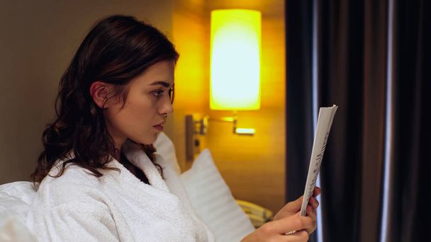 вид сбоку на молодую женщину в халате, читающую газету в гостиничном номере - Фото, изображение