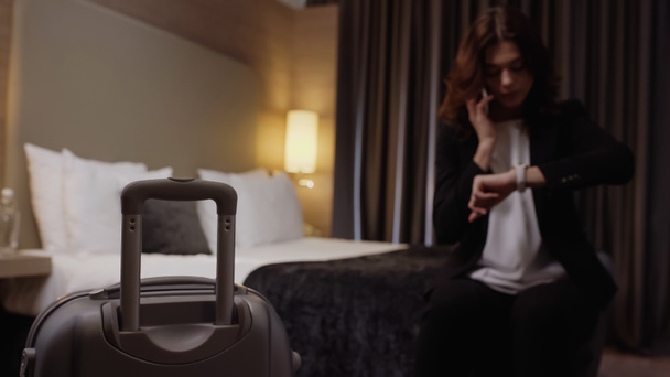 suitcase near blurred businesswoman talking on smartphone and looking at wristwatch in hotel room - Photo, Image