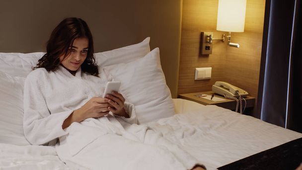 幸せな女性はベッドで休んでいる間にスマートフォンでチャットバスローブで - 写真・画像