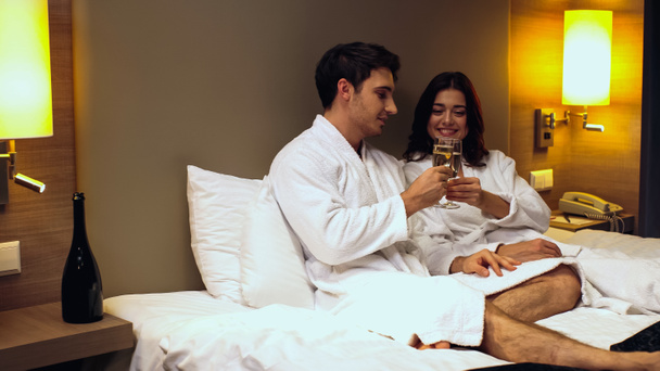 gelukkig paar in badjassen klinkende bril met champagne in hotel kamer - Foto, afbeelding