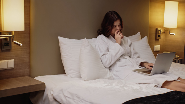 pensive freelancer in bathrobe typing on laptop in bed in hotel room - Foto, afbeelding