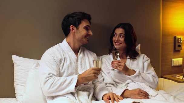 joyful couple in bathrobes holding glasses with champagne in hotel room - Foto, imagen