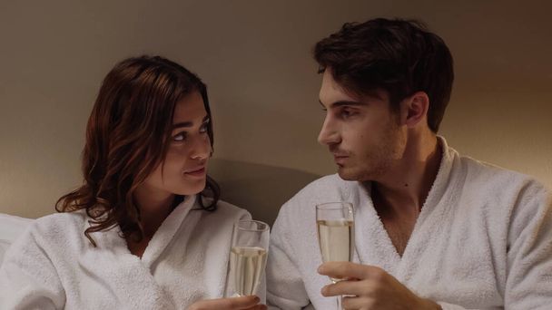 young couple in bathrobes holding glasses with champagne while looking at each other in hotel room - Fotó, kép