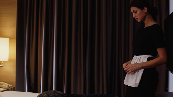 Otel odasında yatağın yanında üniformalı genç bir hizmetçi duruyor. - Fotoğraf, Görsel