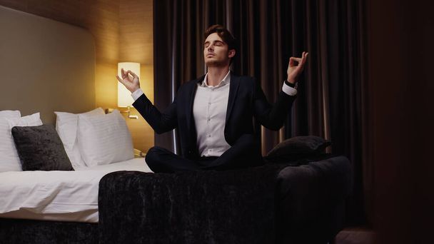 young businessman meditating in lotus pose on bed in hotel room - Photo, Image