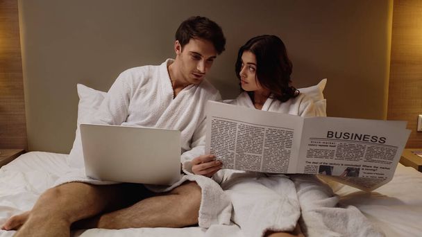 homme avec ordinateur portable lecture journal d'affaires près de jeune copine dans la chambre d'hôtel - Photo, image