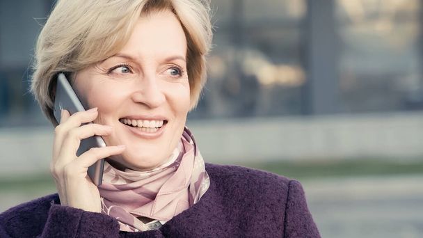 χαρούμενη μεσήλικη γυναίκα κοιτάζει αλλού ενώ μιλάει σε εξωτερικούς χώρους smartphone - Φωτογραφία, εικόνα
